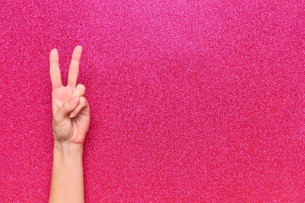 Woman hands making peace and victory symbol on glitter pink background