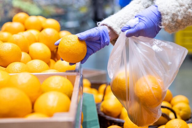 Woman hands in medical gloves chooses fresh oranges folding in disposable plastic bag in supermarket