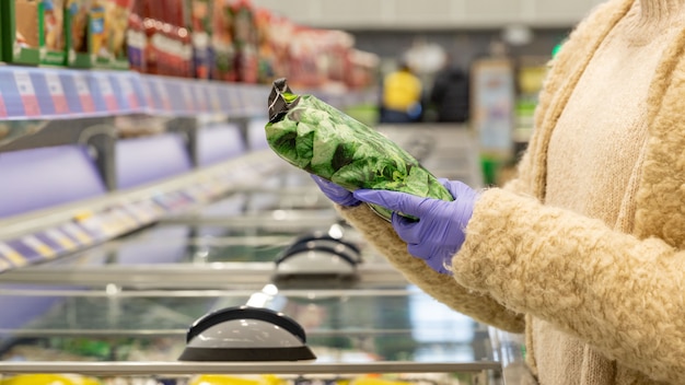 Женщина руки в медицинских перчатках выбирает замороженный шпинат в упаковке, открыв морозильник в супермаркете. Защита от коронавируса