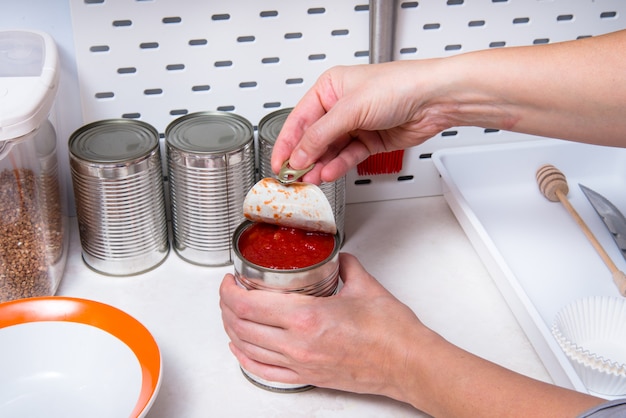 Женщина руки открывая консервную банку с консервированными помидорами, кухонный стол