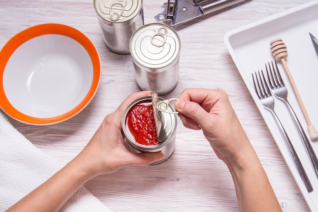 Woman hands opening tin can with canned tomatoes, kitchen table