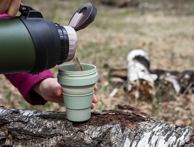 自然の背景に魔法瓶からマグカップにお茶を注ぐ女性の手秋または春の森のトレイルのピットストップサーマルフラスコ