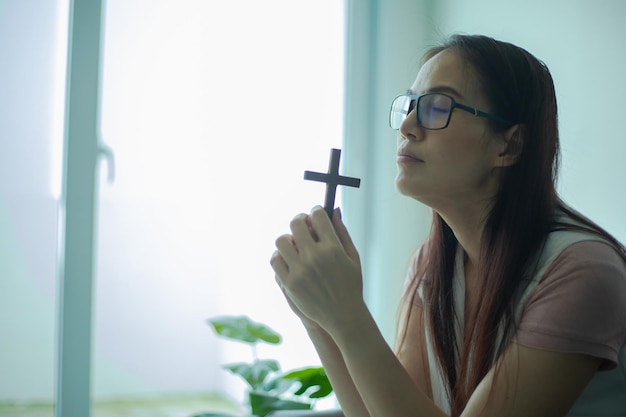 Le mani della donna pregano con una croce di legno vicino alla finestra.