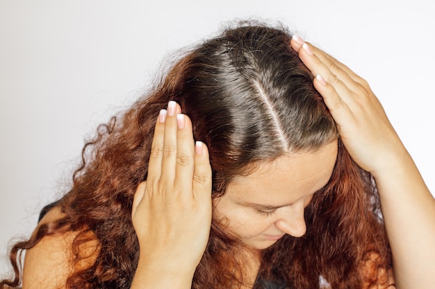 Женщина руками раздвигает волосы в стороны, показывая седеющие корни волос на белом фоне. Отросшие корни, которые нуждаются в перекрашивании. Антивозрастной уход.