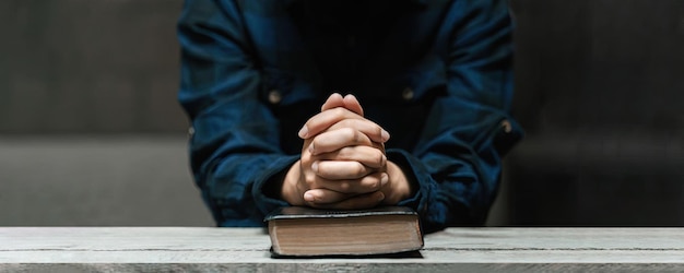 Foto le mani della donna si siedono e pregano con la vecchia bibbia nell'oscurità su un lungo tavolo di legno concetto di amore divino amore fede speranza