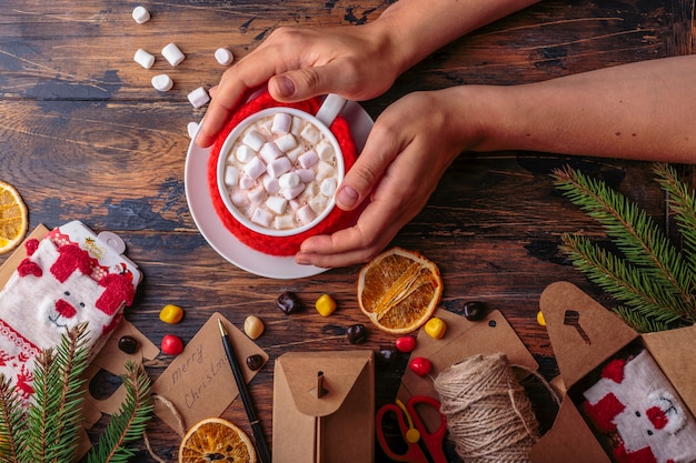 Woman hands takes a mug of hot cacao with marshmallows knitted cloth for a mug christmas concept