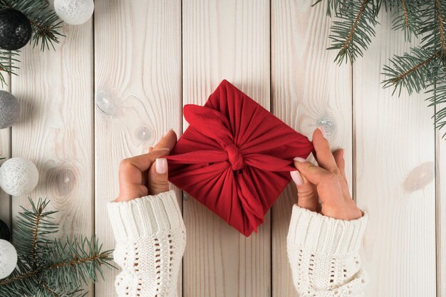 Woman hands in warm sweater tie bow on red furoshiki present box with spruce twigs Christmas toy decoration Top view