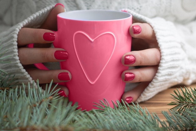Woman hands in white sweater with carmine nail polish hold mug with heart near Christmas tree Color Year 2023 Magenta