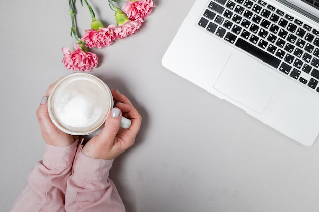 Mani di donna con caffè e laptop fiori garofani su sfondo bianco sfondo dello spazio di lavoro primaverile vista dall'alto