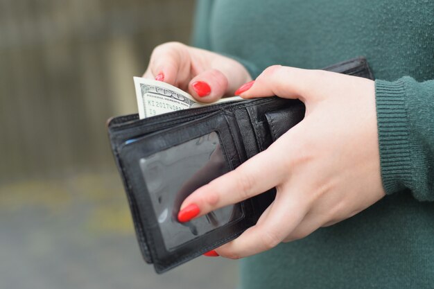 Woman hands with red nails holds black men purse with many US hundred dollars bills