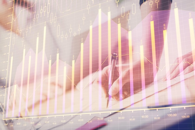 A woman hands writing information about stock market in notepad forex chart holograms in front concept of research double exposure