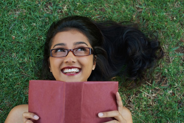 Woman happy and reading book on grass literature and smiling for fiction or fantasy story Female person nature and relaxing on lawn for knowledge textbook and student studying in garden outdoors