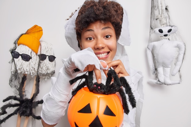 Photo woman has spooky image holds carved pumpkin with big black spider prepares for halloween celebration poses on white believes in mystery awaits for horrible night