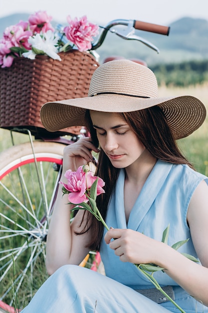 Woman in hat holding pink peony near bicycle with a basket of flowers and enjoys nature. 