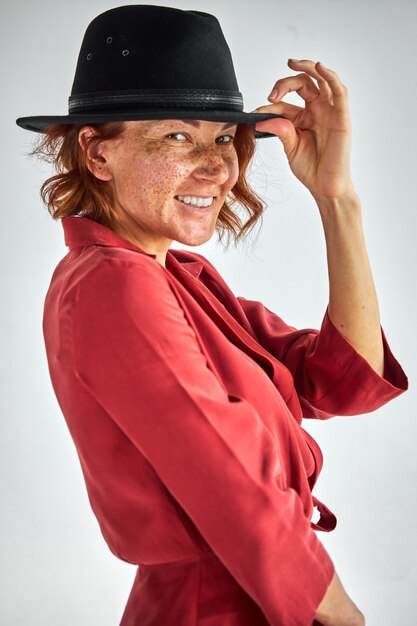 Woman in hat posing at camera, cheerfully smiling, holding hat on head, isolated