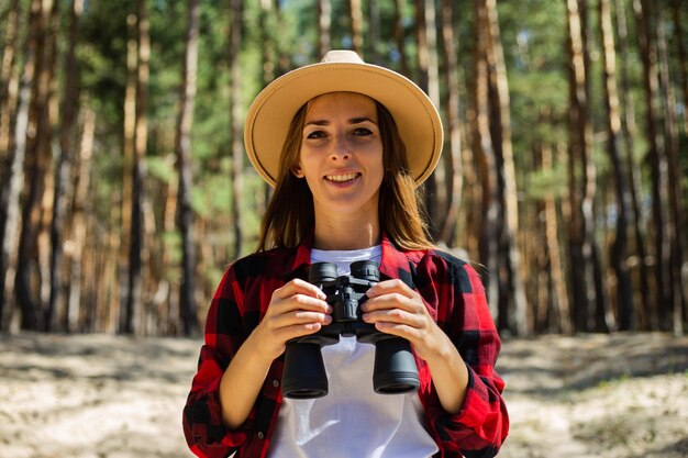 Woman in hat and red plaid shirt holding binoculars in the forest.