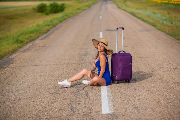 A woman in a hat and with a big bag sits on the pavement Girl in a blue tightfitting dress with a purple suitcase on the roadway A brunette with a sports figure is waiting for a bus