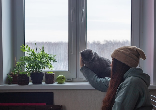 A woman in a hat with ears and long hair is sitting by the window with her cat