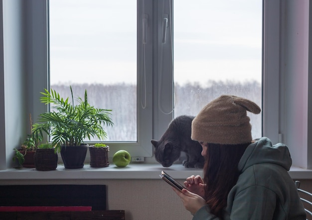 A woman in a hat with ears and long hair is sitting by the window with her cat