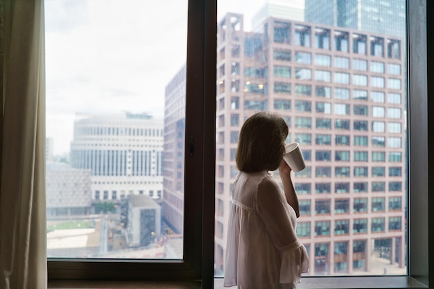 写真 高層ビルの窓から傾いてコーヒーを飲む女性国際コーヒーデーのコンセプト