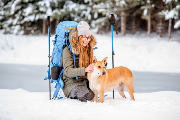 湖と森の近くの雪山で犬をなでる冬のハイキング中に休憩をとっている女性