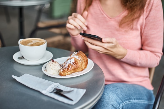 Donna che mangia prima colazione in un caffè e che utilizza smartphone