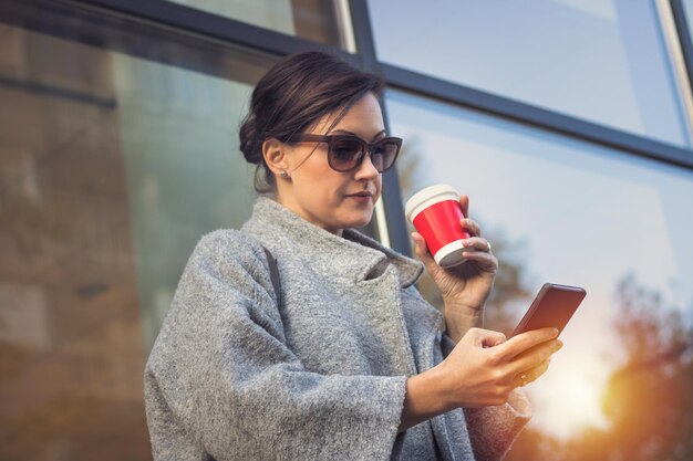 Foto donna che beve caffè mentre usa il cellulare in città