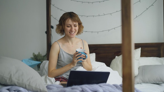 woman having online video chat with friends using laptop camera while sitting on bed at home