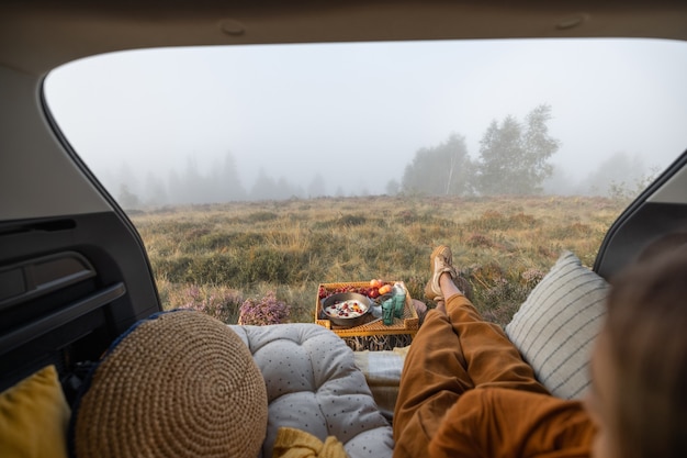 Photo woman having a picnic and enjoying great view on nature in foggy weather, sitting relaxed in the vehicle trunk, view from a car interior