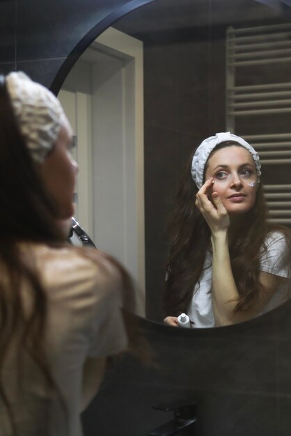 A woman in a headband applies moistening cream for eye skin in front of a mirror Natural skincare Loft interior bathroom Beauty home spa