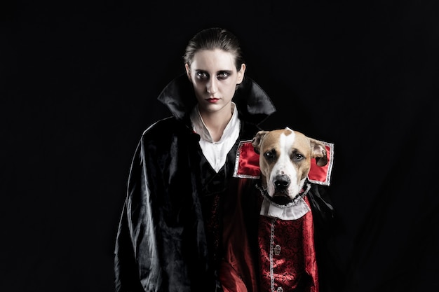 Woman and her dog in similar vampire costumes for halloween. Young female and her pet puppy dressed up in matching dracula costume, posing in black studio background