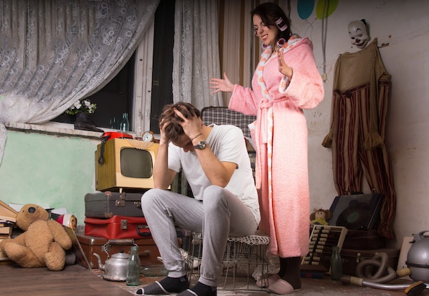 Woman in her dressing gown and slippers standing nagging her husband who is covering his ears in a squalid untidy living room