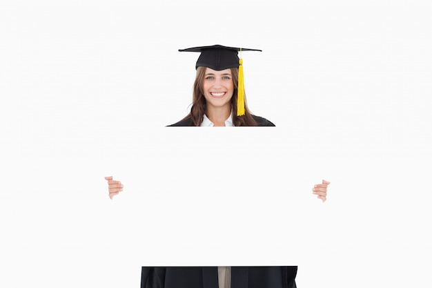 A woman in her graduation gown holds a blank sheet in front of her