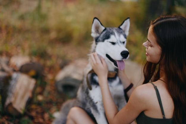 女性と彼女のハスキー犬は、公園の木々の間で屋外で楽しく遊んでおり、秋の散歩にはペットと一緒に歯を見せて微笑んでいます