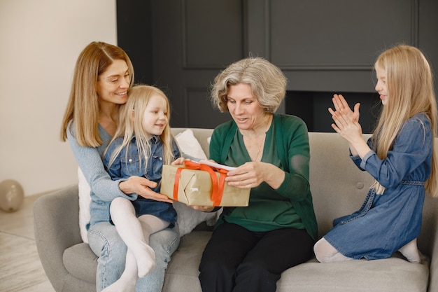 Donna, sua madre e sua figlia sedute su un divano. la ragazza tiene una scatola con un regalo e la dà a sua nonna.