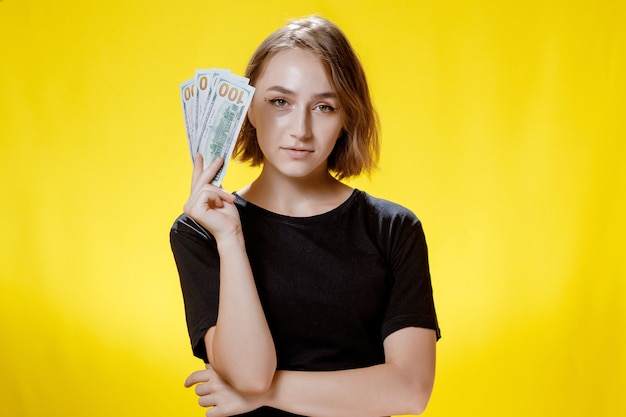 Woman hiding behind bunch of money banknotes and celebrating over yellow background.