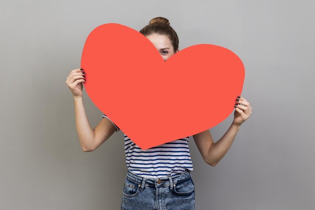 Woman hiding face behind big red heart and looking at camera with curious prying eyes