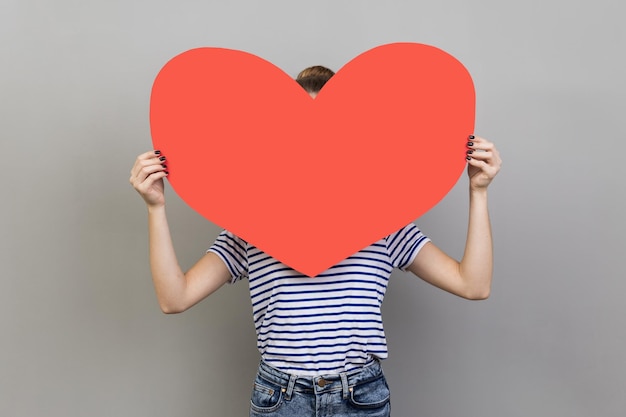 Woman hiding her face behind big red heart hesitates to confess\
his feelings