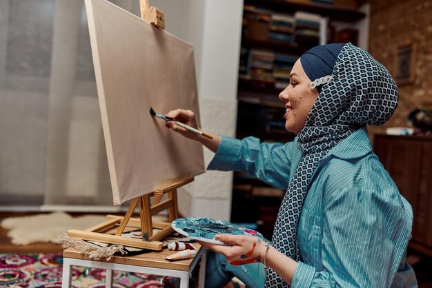 A woman in a hijab paints on canvas with a brush and tempera