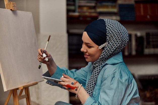 A woman in a hijab paints on canvas with a brush and tempera