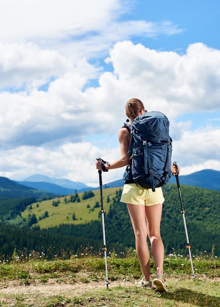 Woman hiker hiking on grassy hill, wearing backpack, using trekking sticks in the mountains