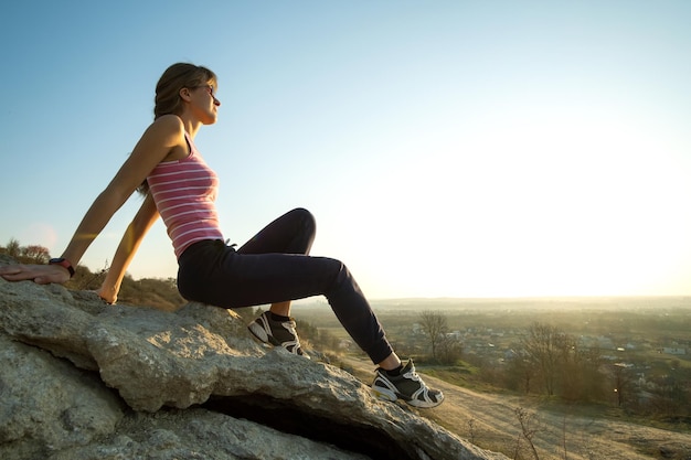 Woman hiker sitting on a steep big rock enjoying warm summer day Young female climber resting during sports activity in nature Active recreation in nature concept