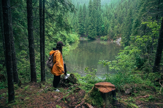 Woman hiker with backpack in yellow raincoat looking at mountain lake