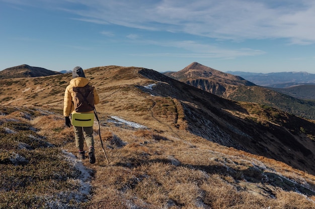 Woman hiker in yellow jacket with backpack climbs autumn
mountains with light snow on ground on sunn