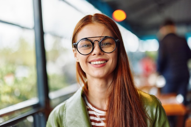 Woman hipster with red hair and glasses sits in town in cafe smile with teeth looking at camera woman freelance blogger closeup