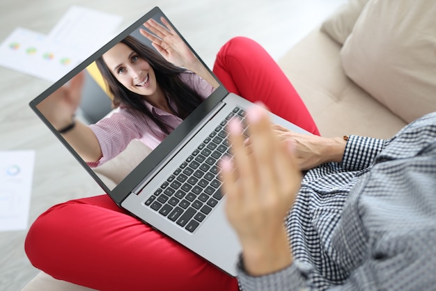 Woman hold computer on her lap and wave hello. On laptop screen cheerful woman