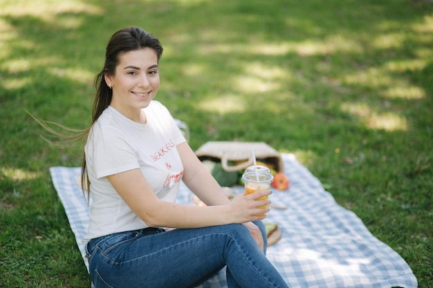 Woman hold disposable cup with orange lemonade on picnic outdoors Space for text Vegan picnic concept