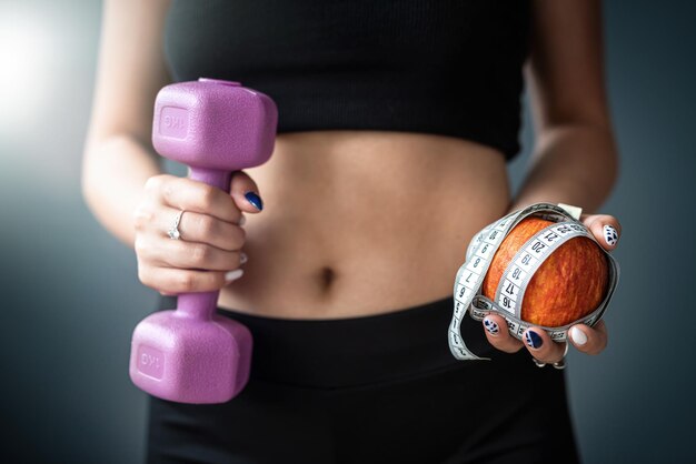 Woman in holding apple and measuring tape and dumbbell standing isolated over gray background