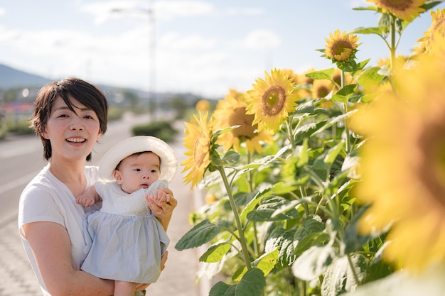 A woman holding a baby and wearing a hat stands in front of a sunflower field.