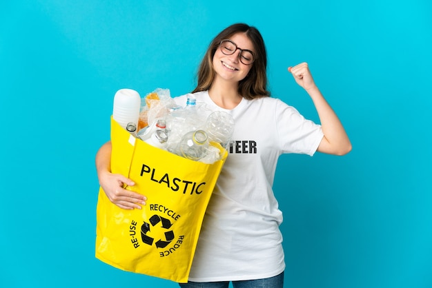 Woman holding a bag full of bottles to recycle isolated on blue doing strong gesture